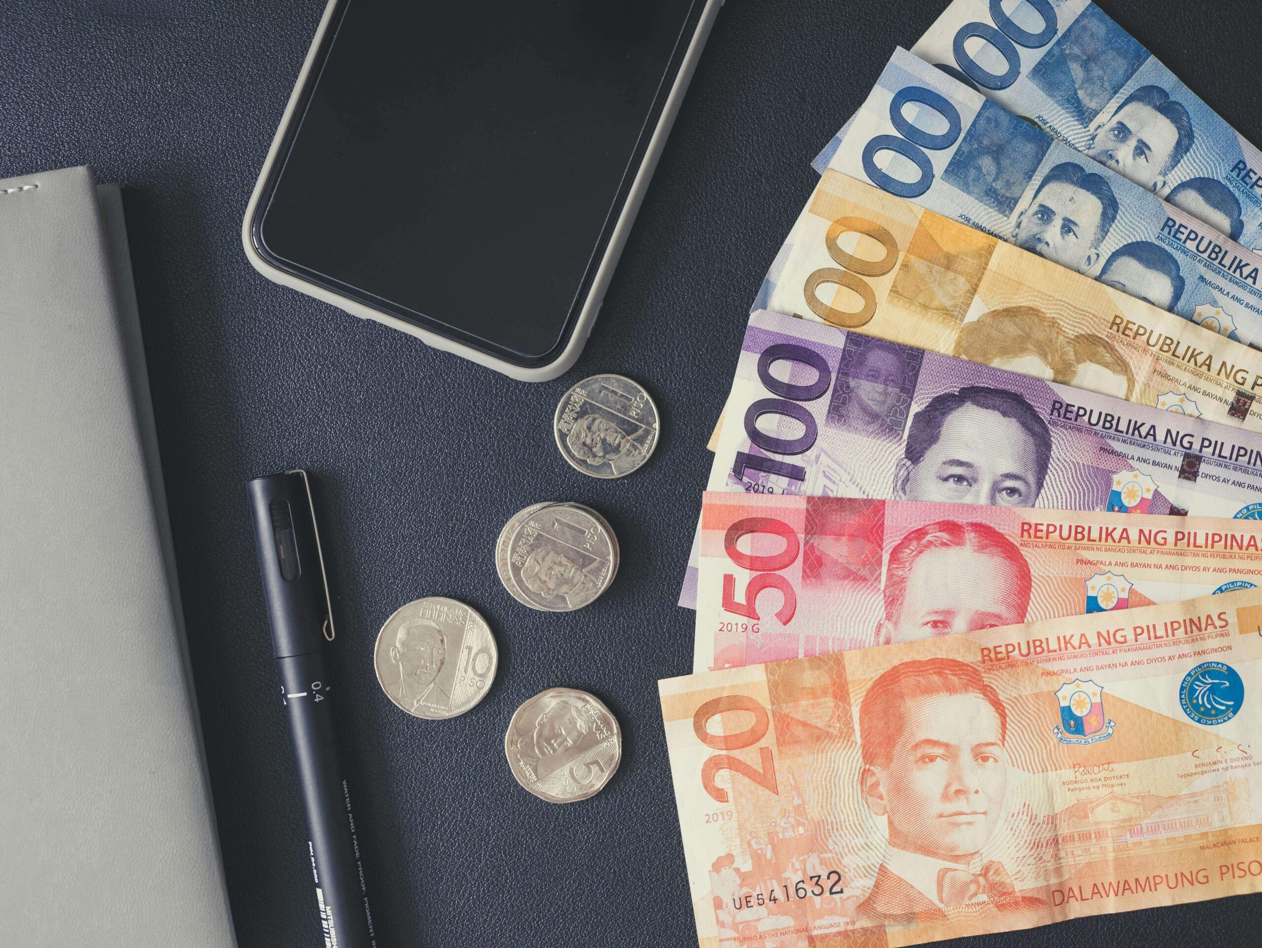 Philippine peso paper bills and coins displayed with a phone and a pen